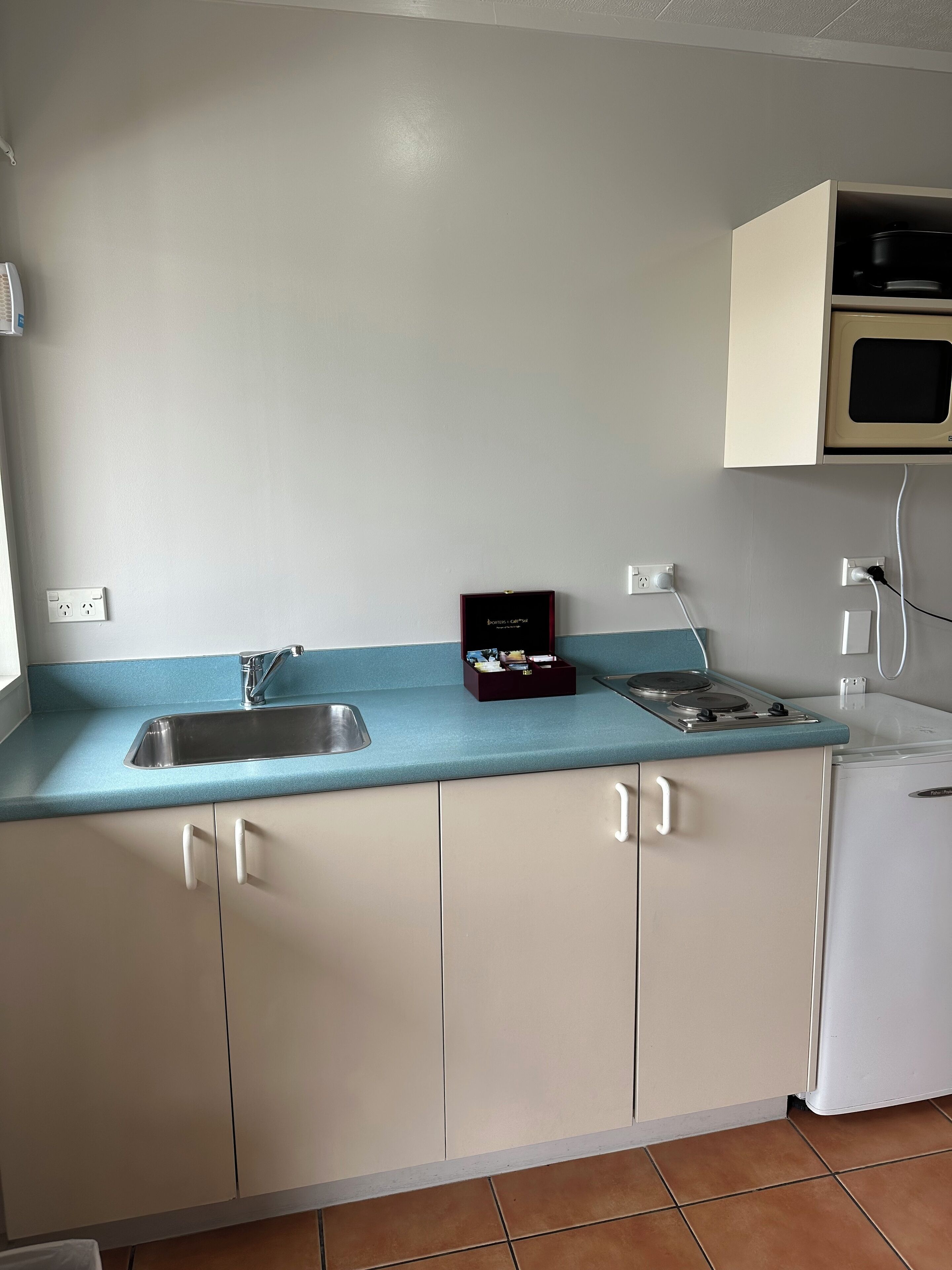 A compact kitchen with a sink, stove, and microwave. There is a small cabinet with a dark-colored box on the counter.