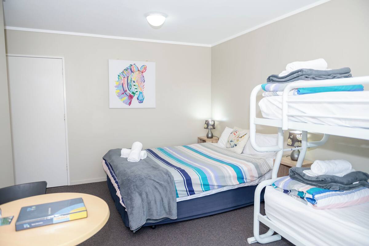 Habitación de hotel con una litera, una cama individual, un escritorio con silla y un cuadro en la pared.