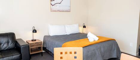 A hotel room with a bed, a black leather chair, a wooden nightstand, a table with a remote control, and a painting on the wall.