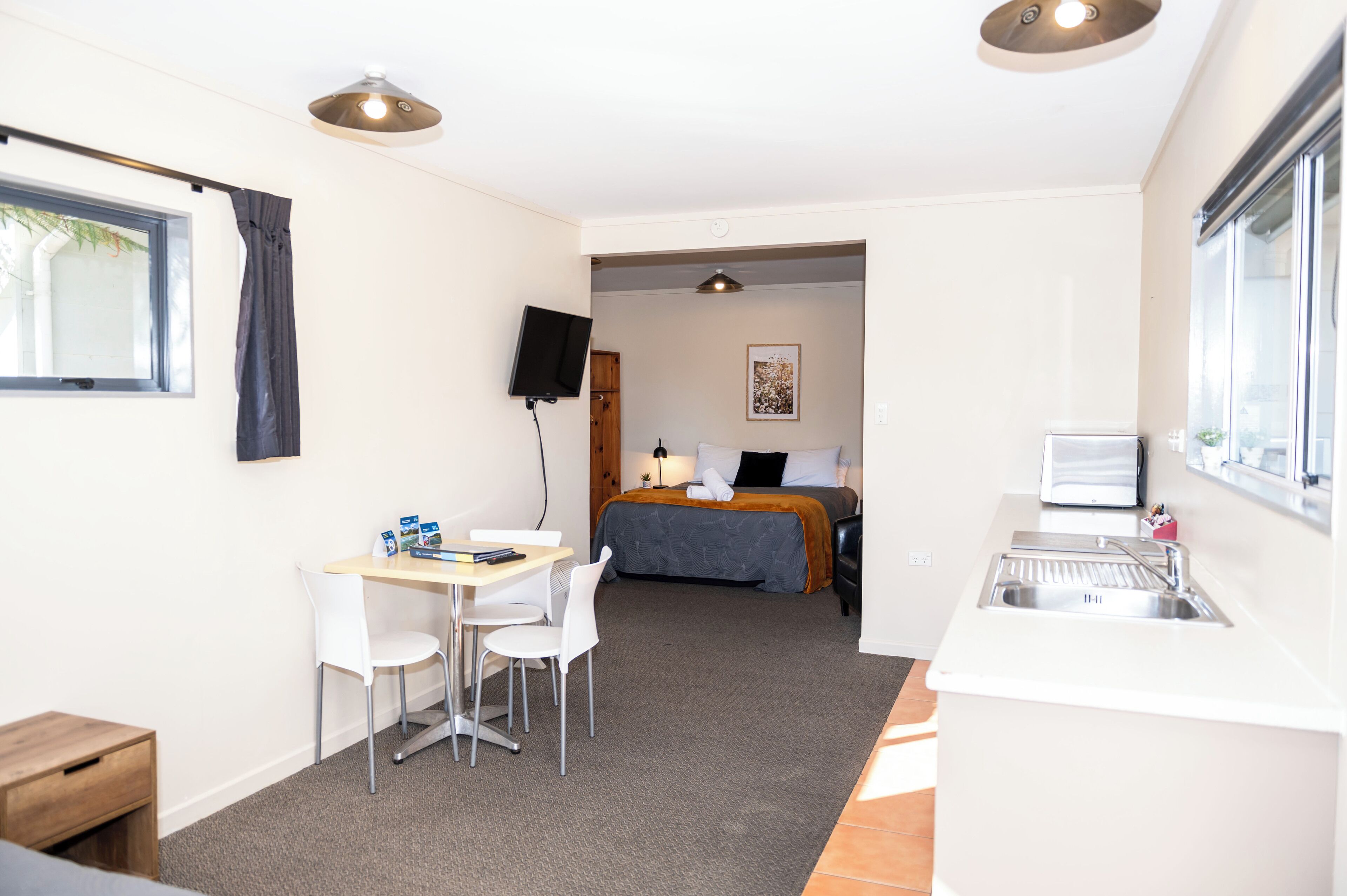 A compact hotel room with a kitchenette, a bed with a gray blanket, a desk with a chair, and a TV mounted on the wall.