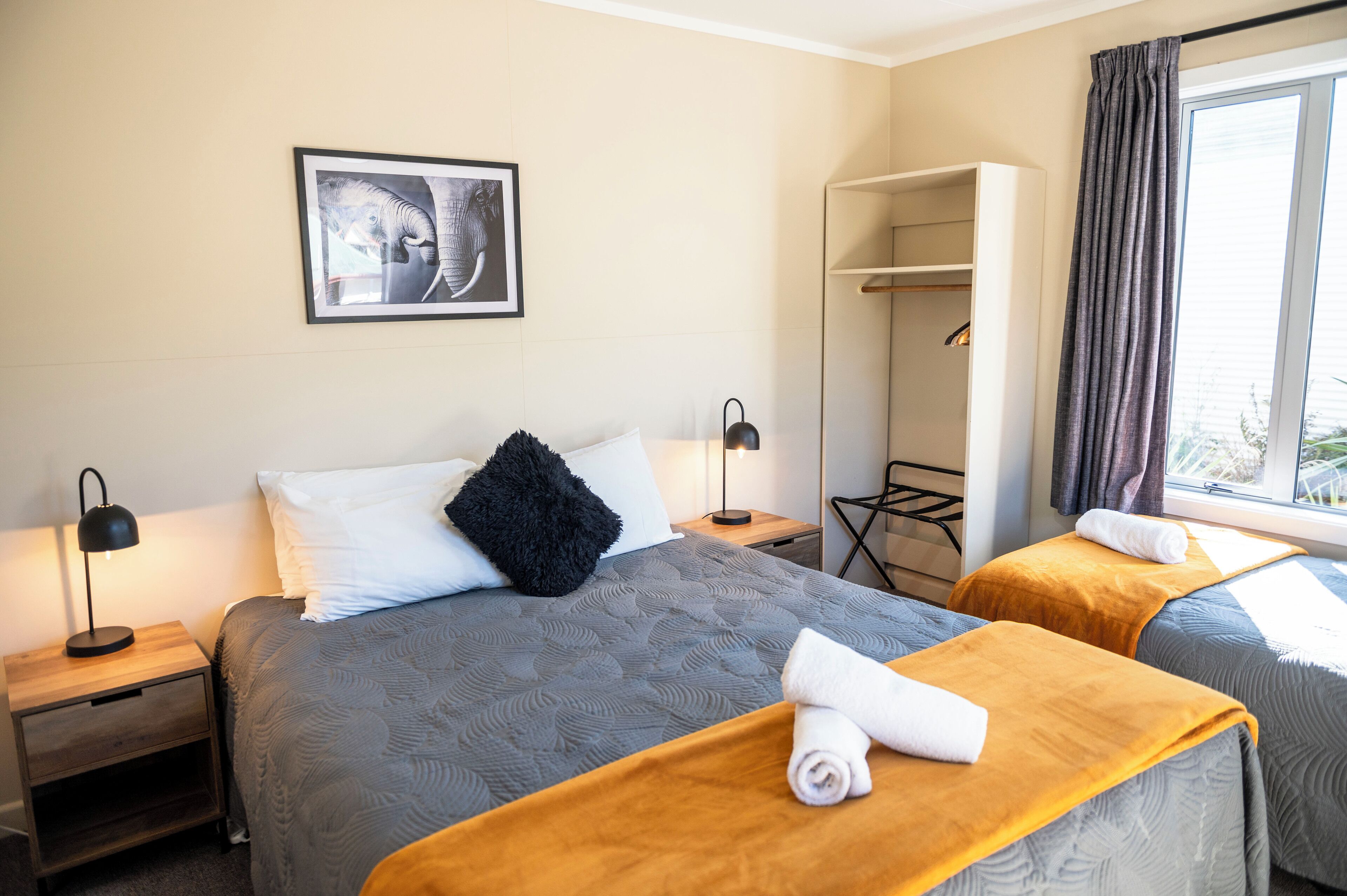Habitación de hotel con dos camas, una mesita de noche con lámpara, un armario y una ventana con cortinas.