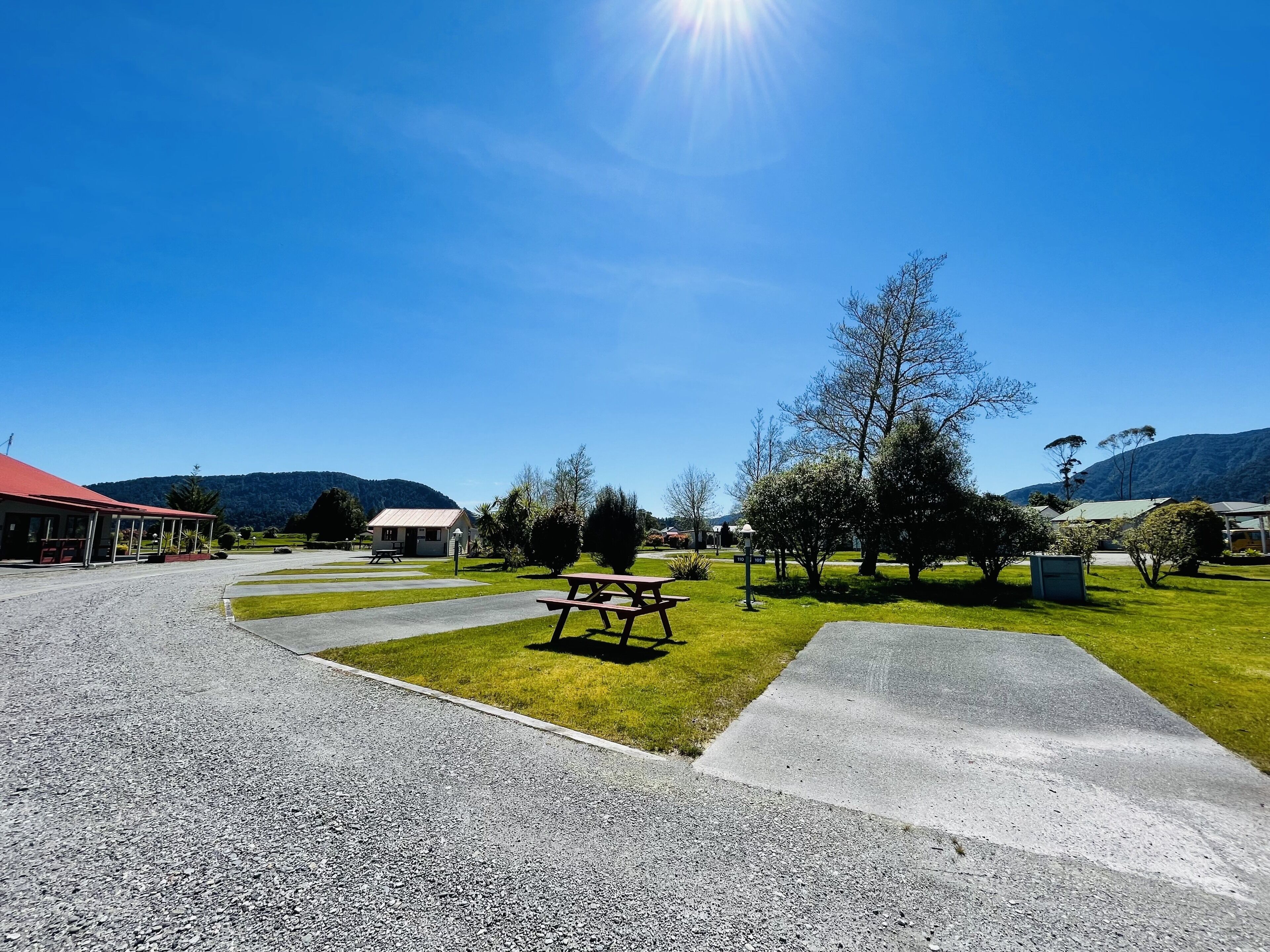 A picnic area with a bench and picnic table, surrounded by grass and trees, with a building in the background.