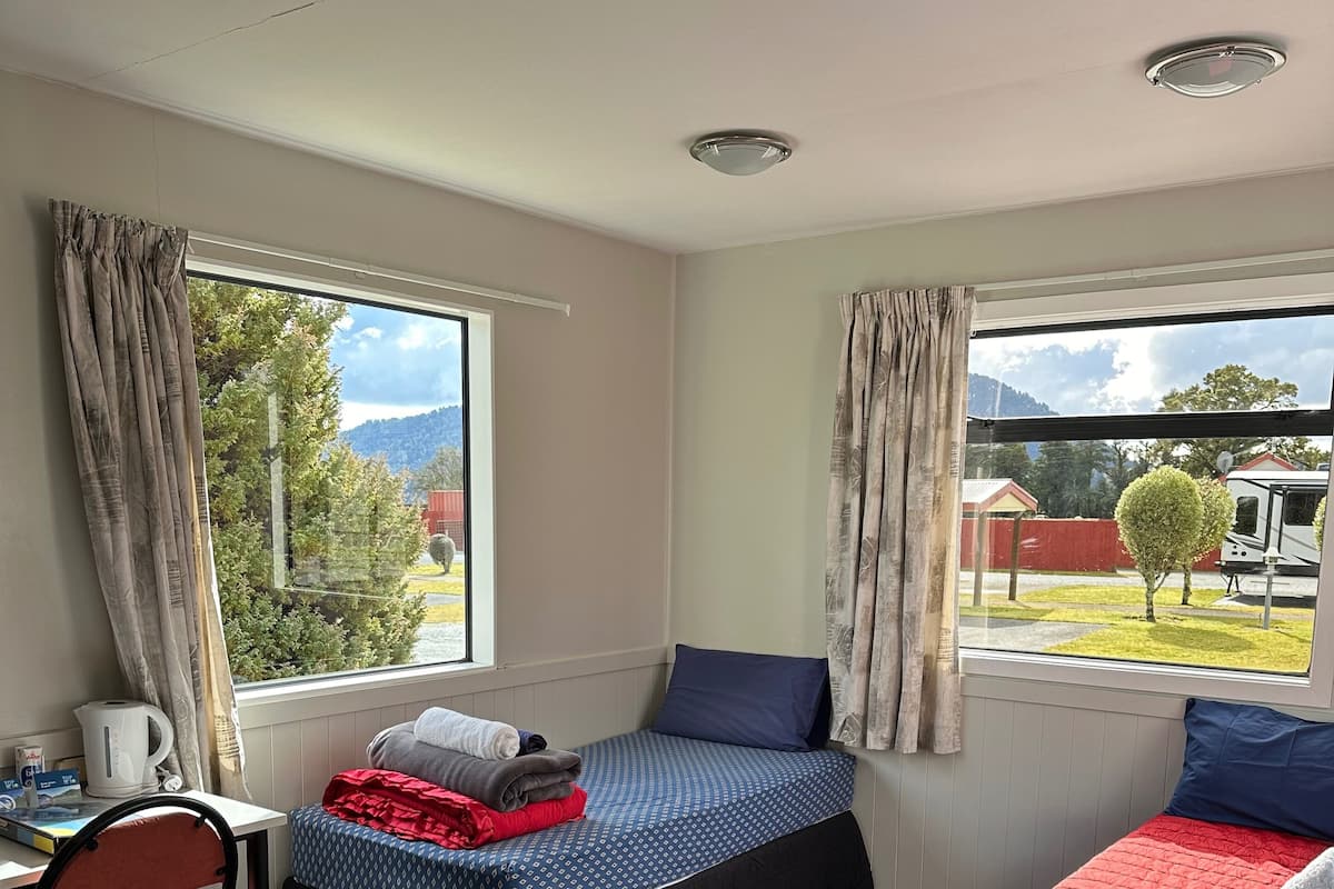 Una habitación con dos camas, un escritorio y una silla. Hay ventanas con cortinas, vistas al exterior y una grieta en el techo.