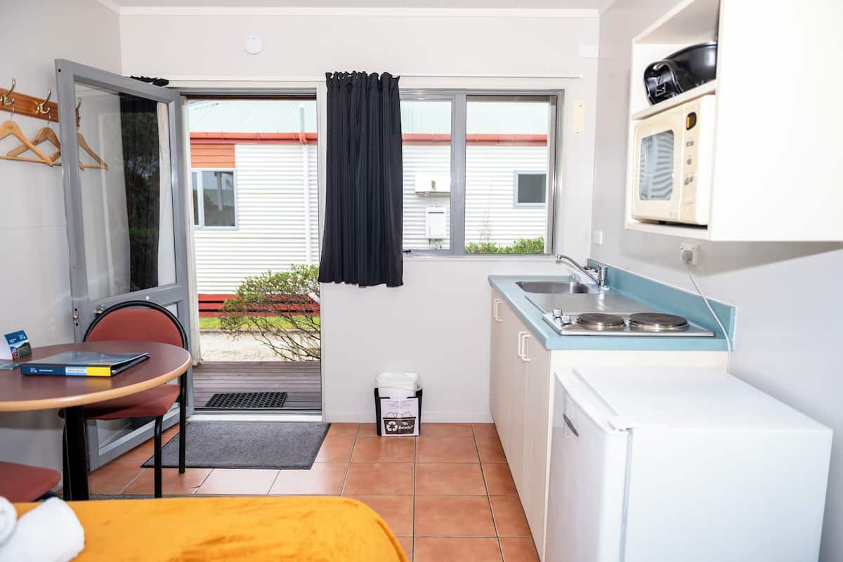 Cocina compacta con lavamanos, microondas y un refrigerador pequeño. Hay un comedor con una mesa y sillas, y una ventana con cortina.