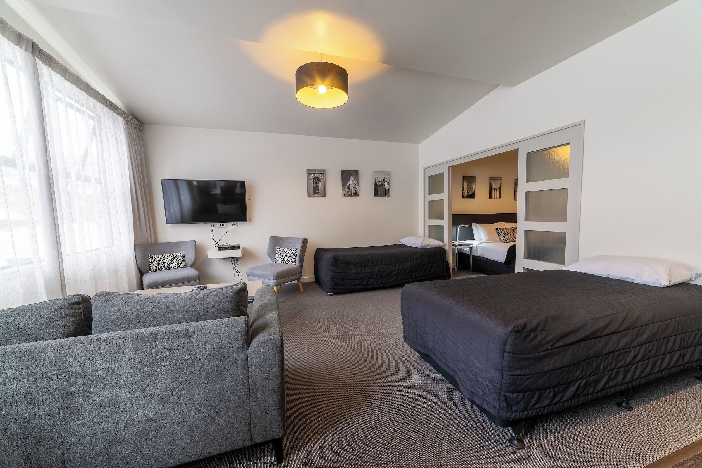 Una habitación de hotel moderna con sofá, dos camas individuales, televisor y un ventanal con cortinas.