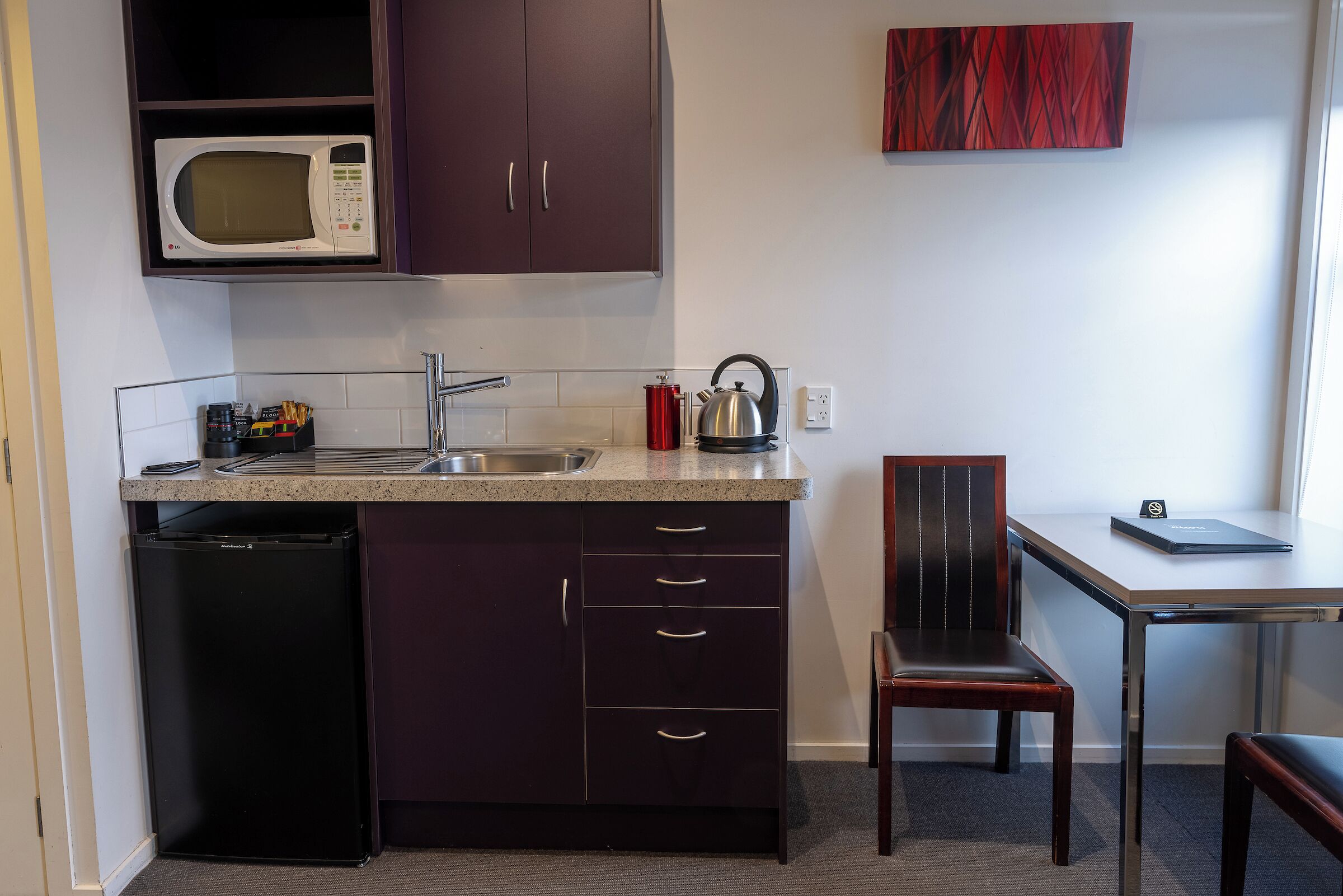 A compact kitchen with a microwave, sink, and a small refrigerator. There's a chair and a table with a book on it.