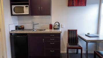 A compact kitchen with a microwave, sink, and a small refrigerator. There's a chair and a table with a book on it.