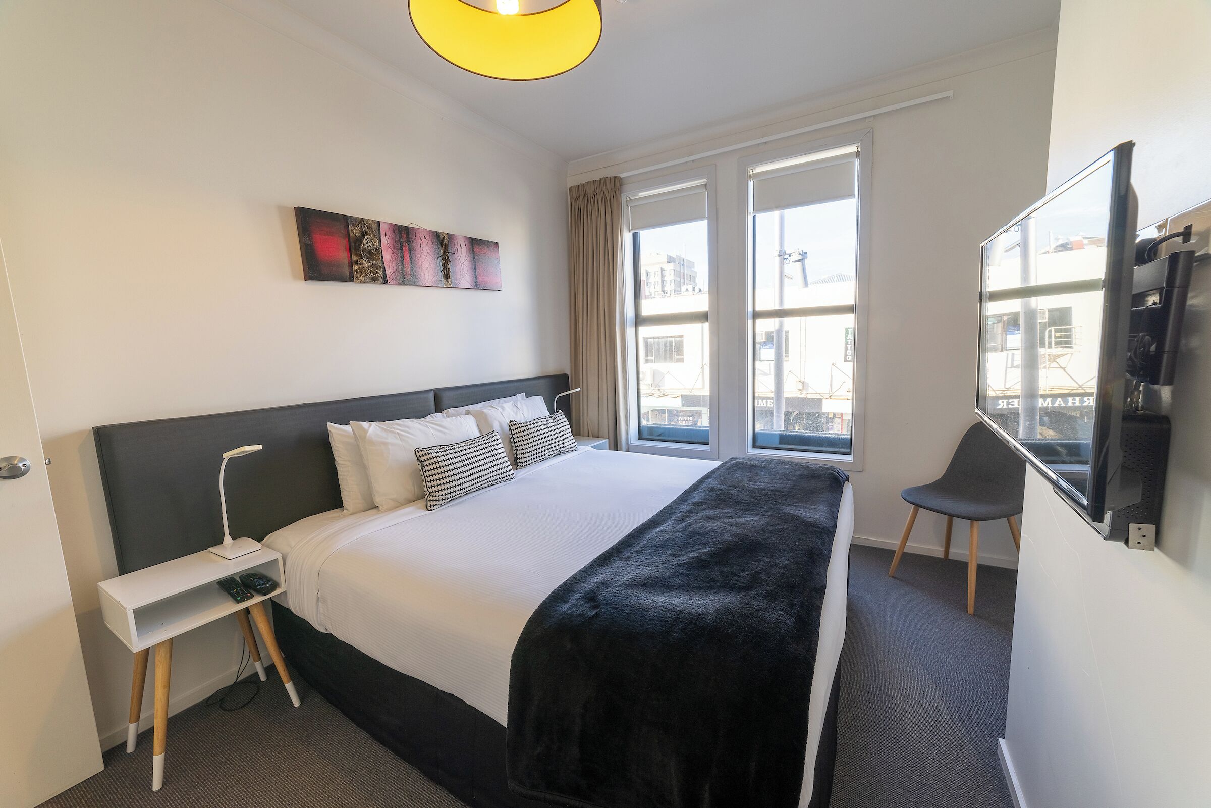 Habitación de hotel con una cama grande, una mesita de noche con lámpara, una silla y un televisor montado en la pared.