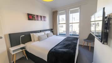 Habitación de hotel con una cama grande, una mesita de noche con lámpara, una silla y un televisor montado en la pared.