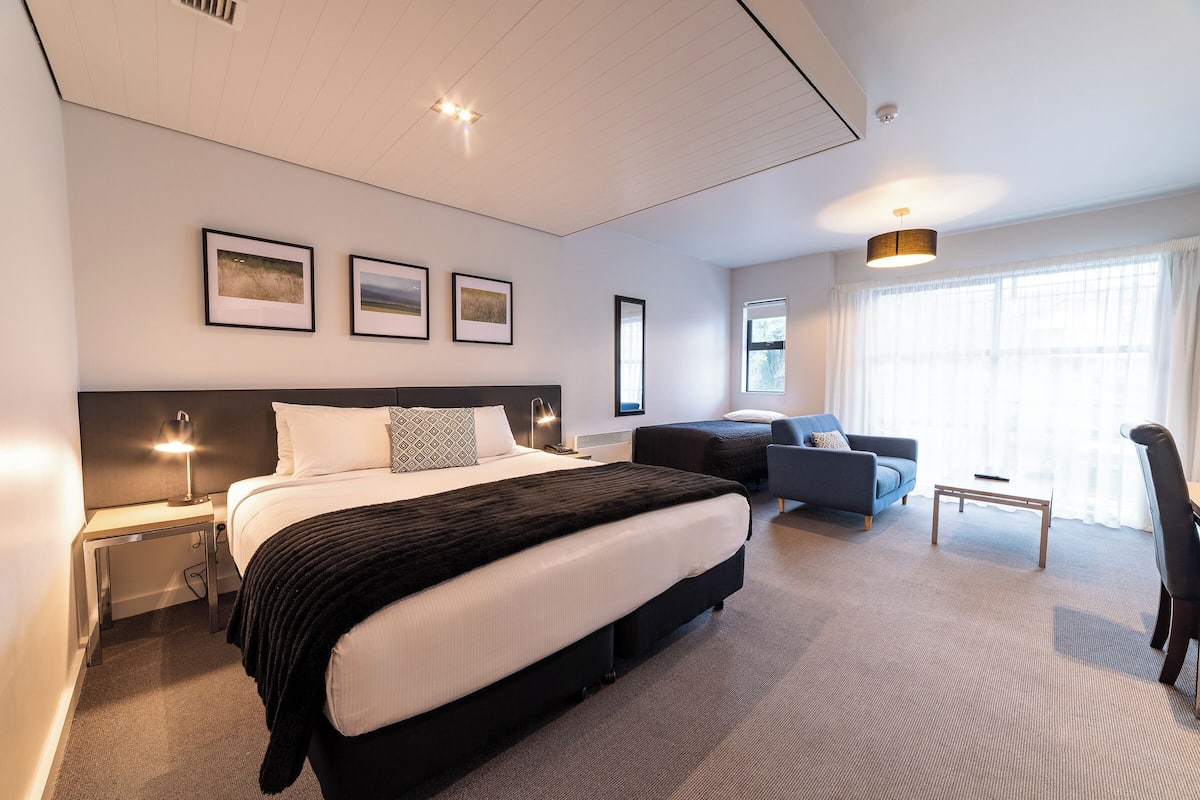 A modern hotel room with a large bed, a desk, a sofa, and a chair.