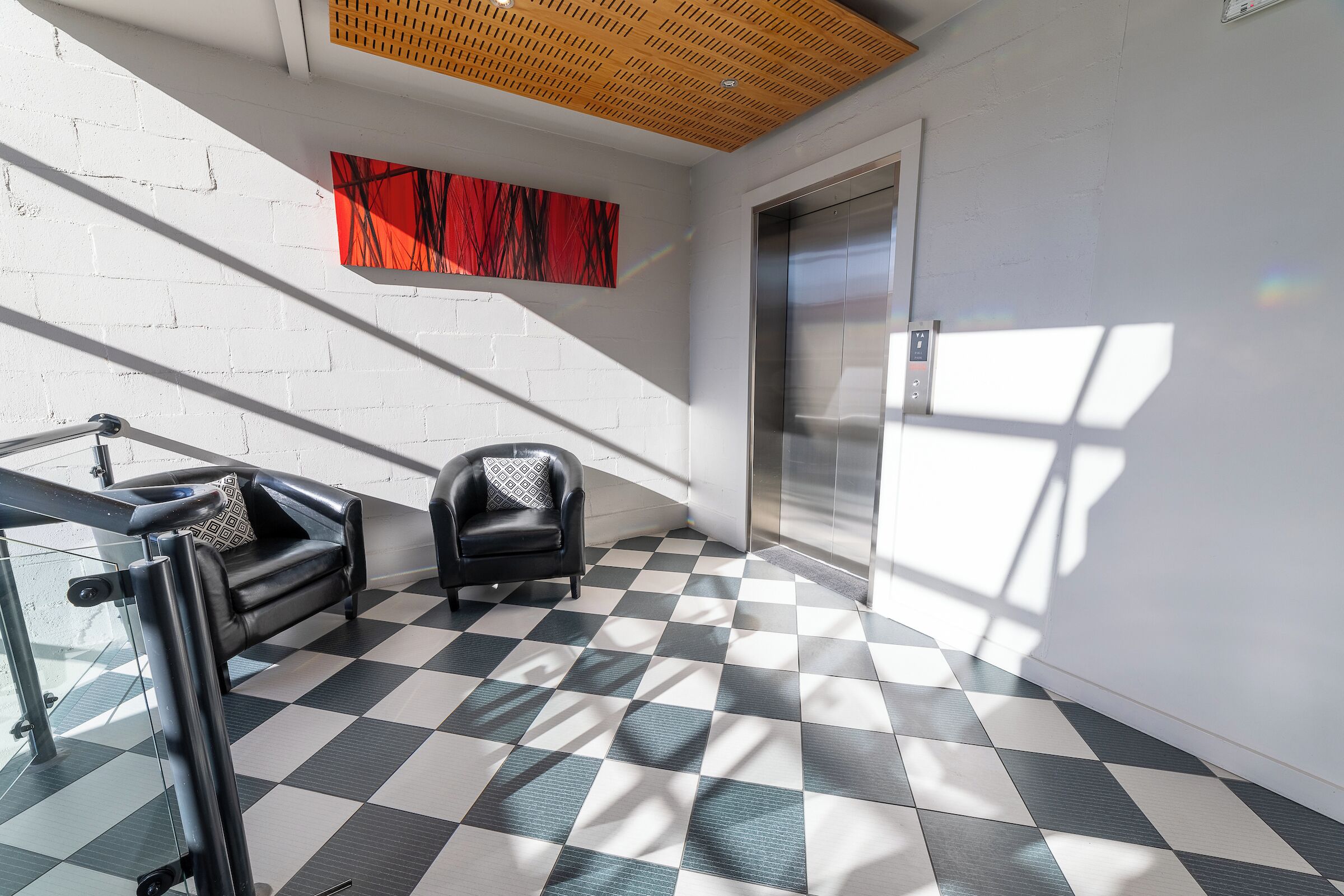 A modern lobby with a checkered floor, a staircase, a black leather sofa, and a red abstract painting on the wall.