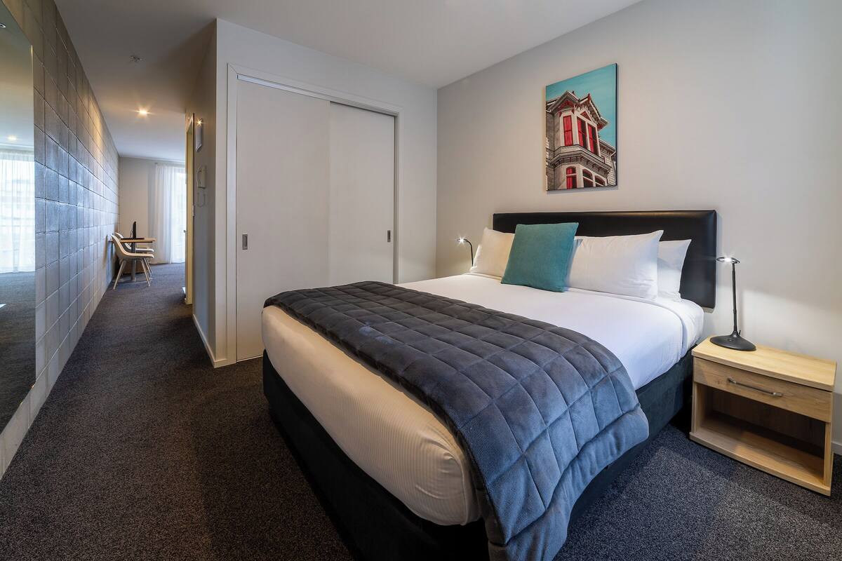 A modern hotel room with a large bed, a bedside table, a nightstand, and a painting on the wall.