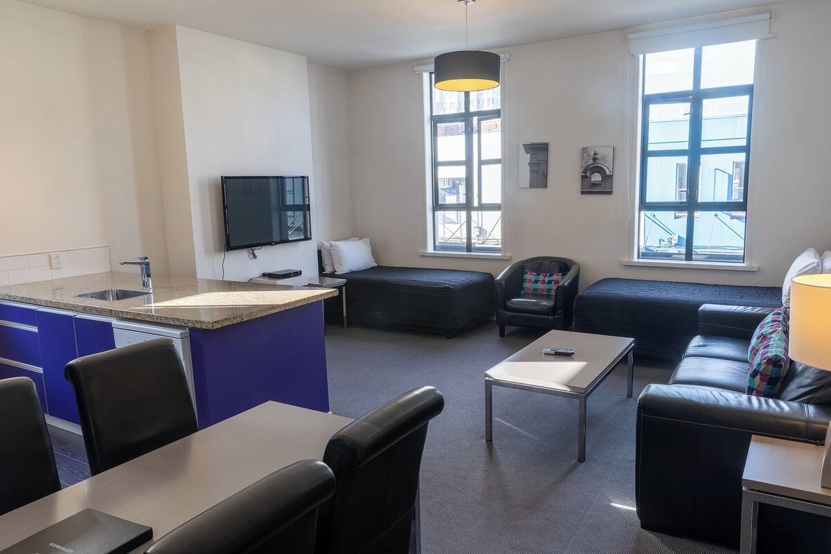 A modern living room with a sofa, a coffee table, and a TV. There is a kitchen area with a sink and cabinets.