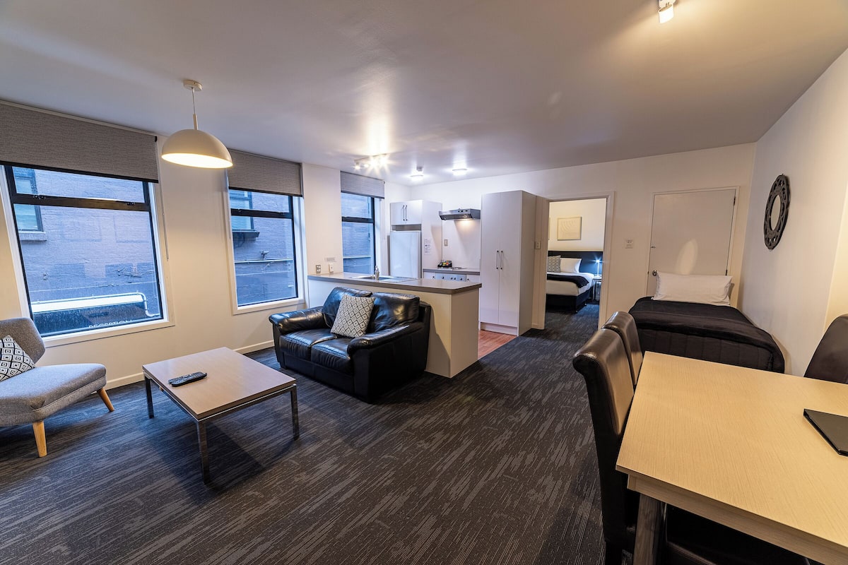 A modern living room with a sofa, coffee table, and a dining table. There is a kitchen area with cabinets and a refrigerator. The room has large windows and a ceiling light fixture.