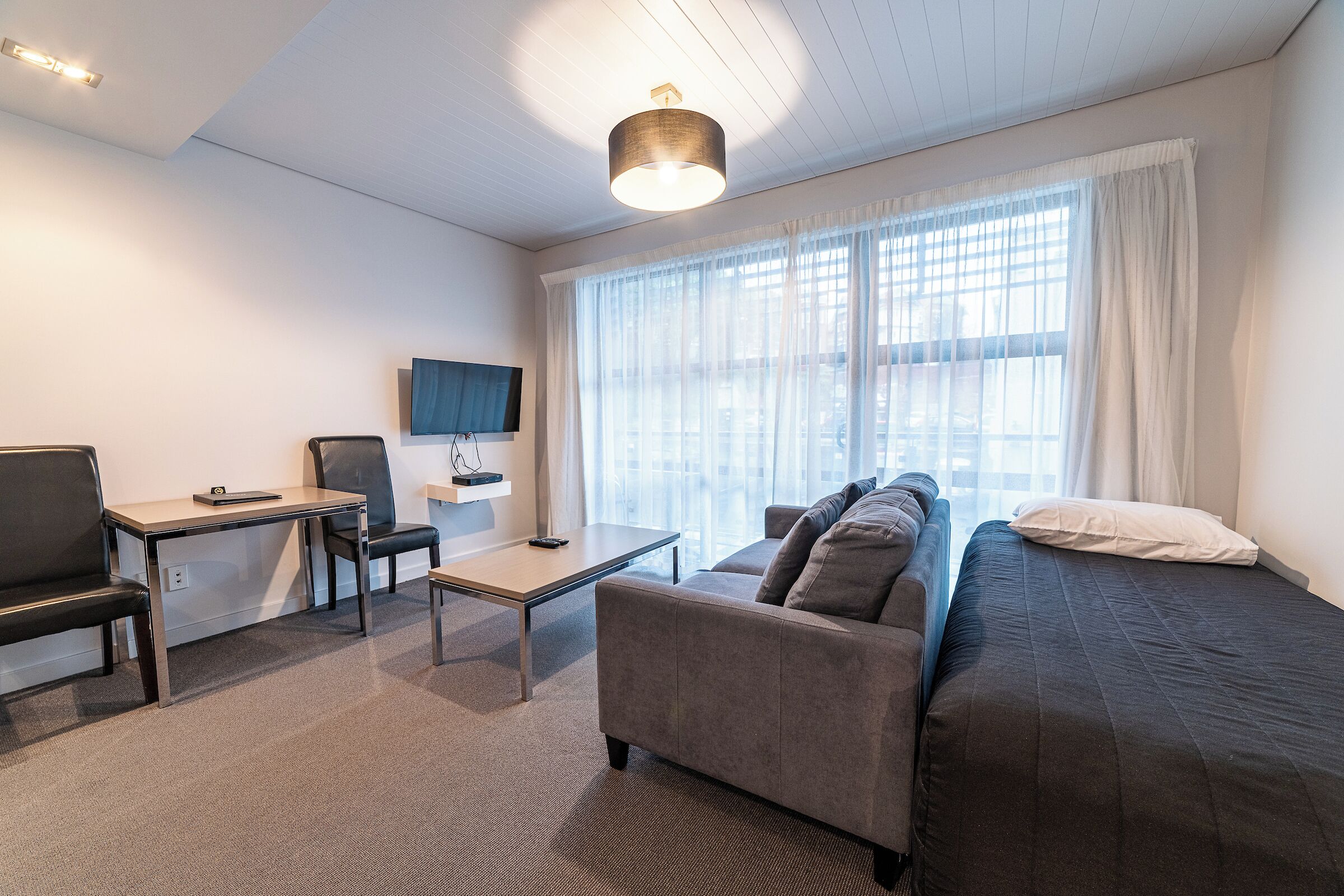 A modern hotel room with a sofa, a desk, a chair, a TV, and large windows with sheer curtains.