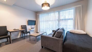 A modern hotel room with a sofa, a desk, a chair, a TV, and large windows with sheer curtains.