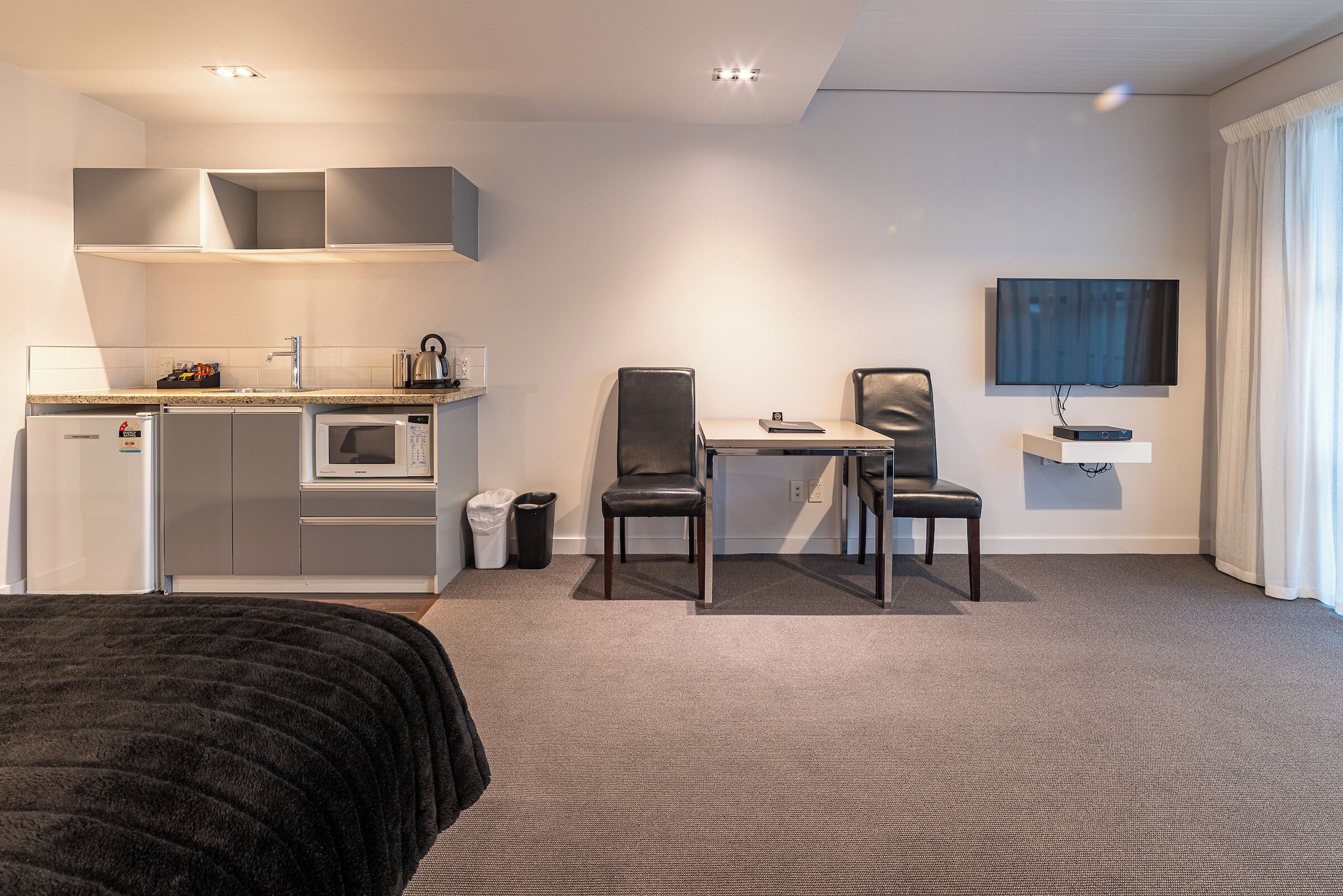 A modern hotel room with a kitchenette, a dining area with two chairs, a TV mounted on the wall, and a large window with curtains.