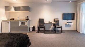A modern hotel room with a kitchenette, a dining area with two chairs, a TV mounted on the wall, and a large window with curtains.