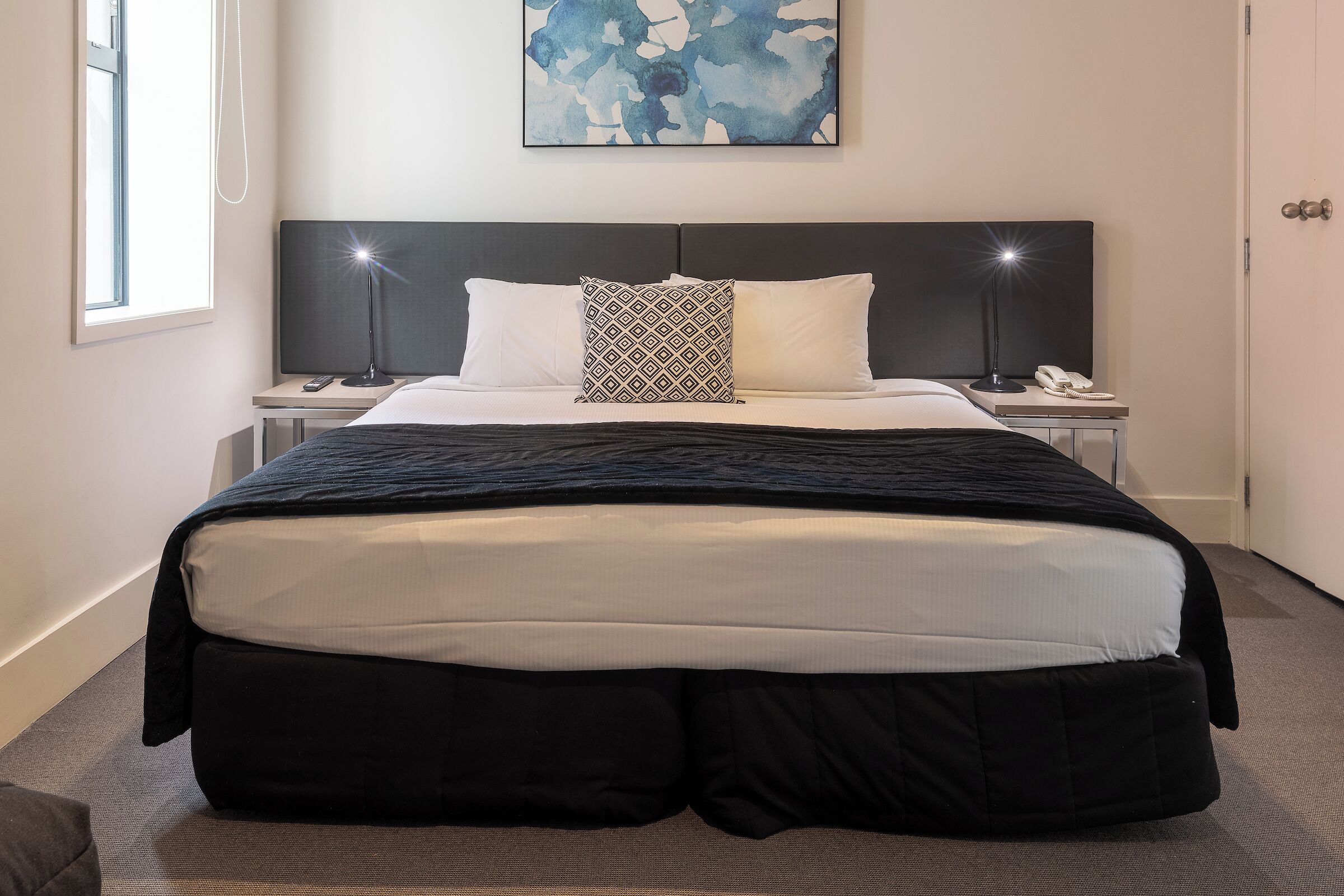 Una cama bien hecha con cabecera negra y blanca, dos lámparas de noche y un cuadro abstracto enmarcado sobre la cama.