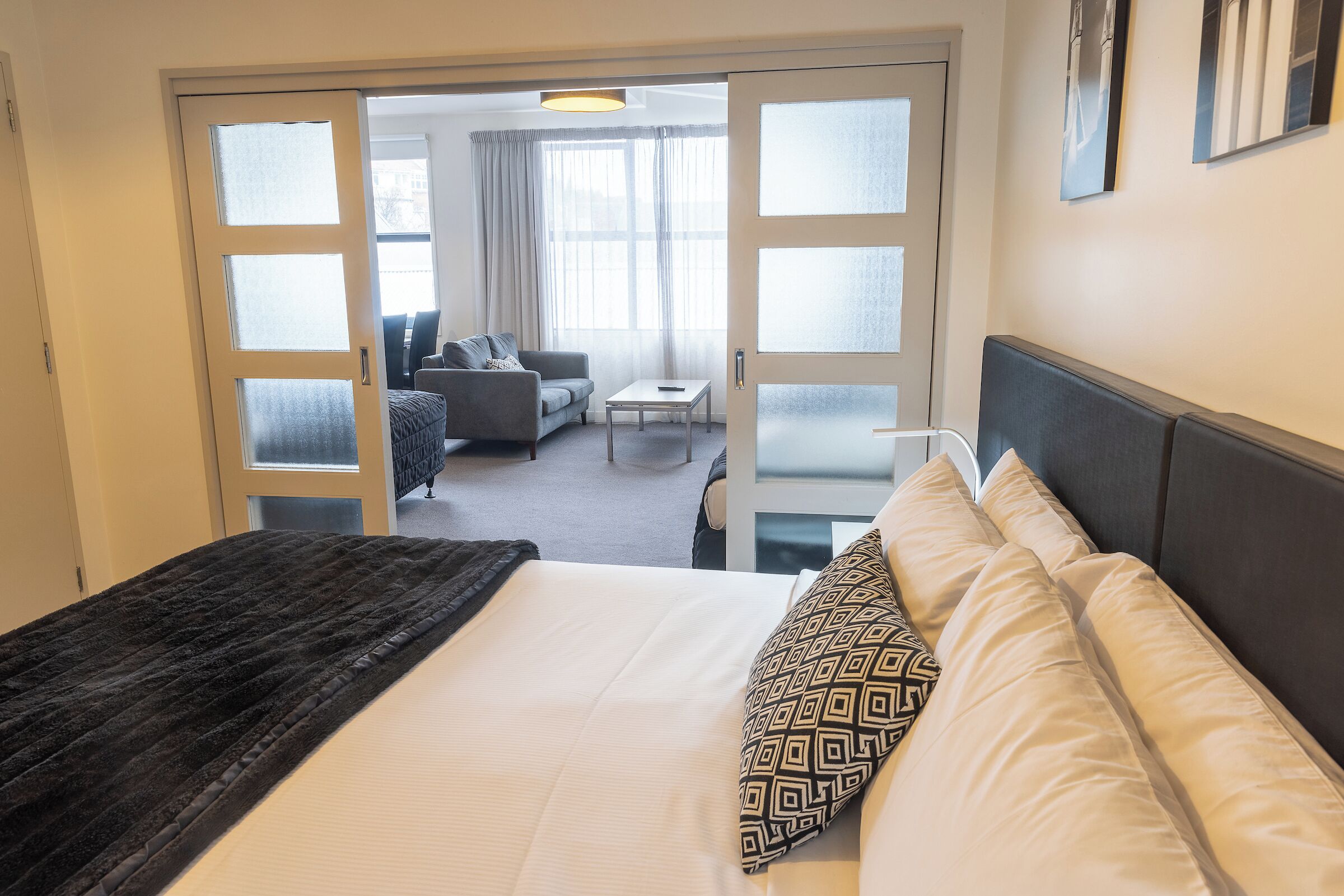 A hotel room with a large bed, a dark throw blanket, patterned pillows, and a view of the adjacent living area through a glass door.