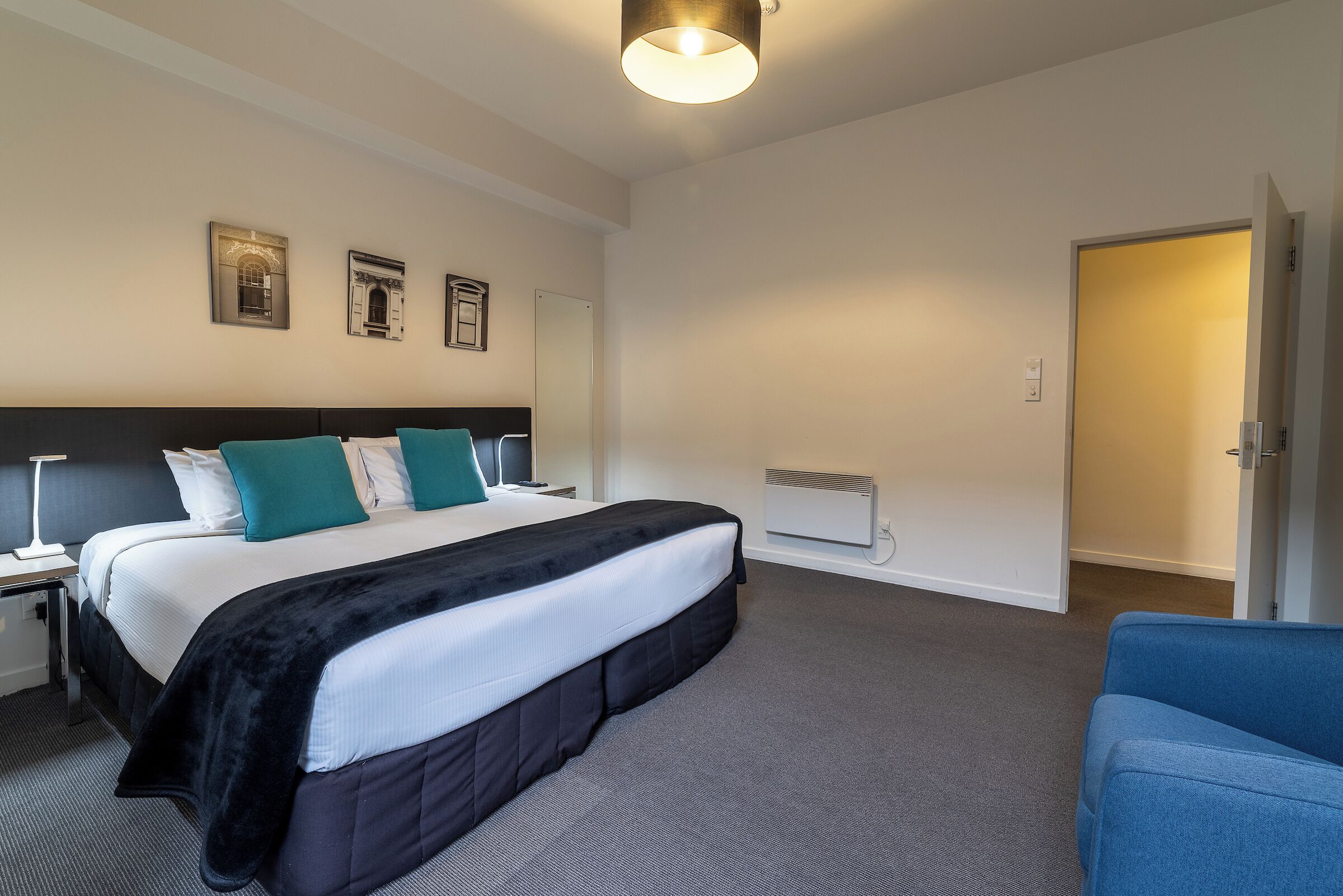 A hotel room with a large bed, a desk with a lamp, a chair, and a door.