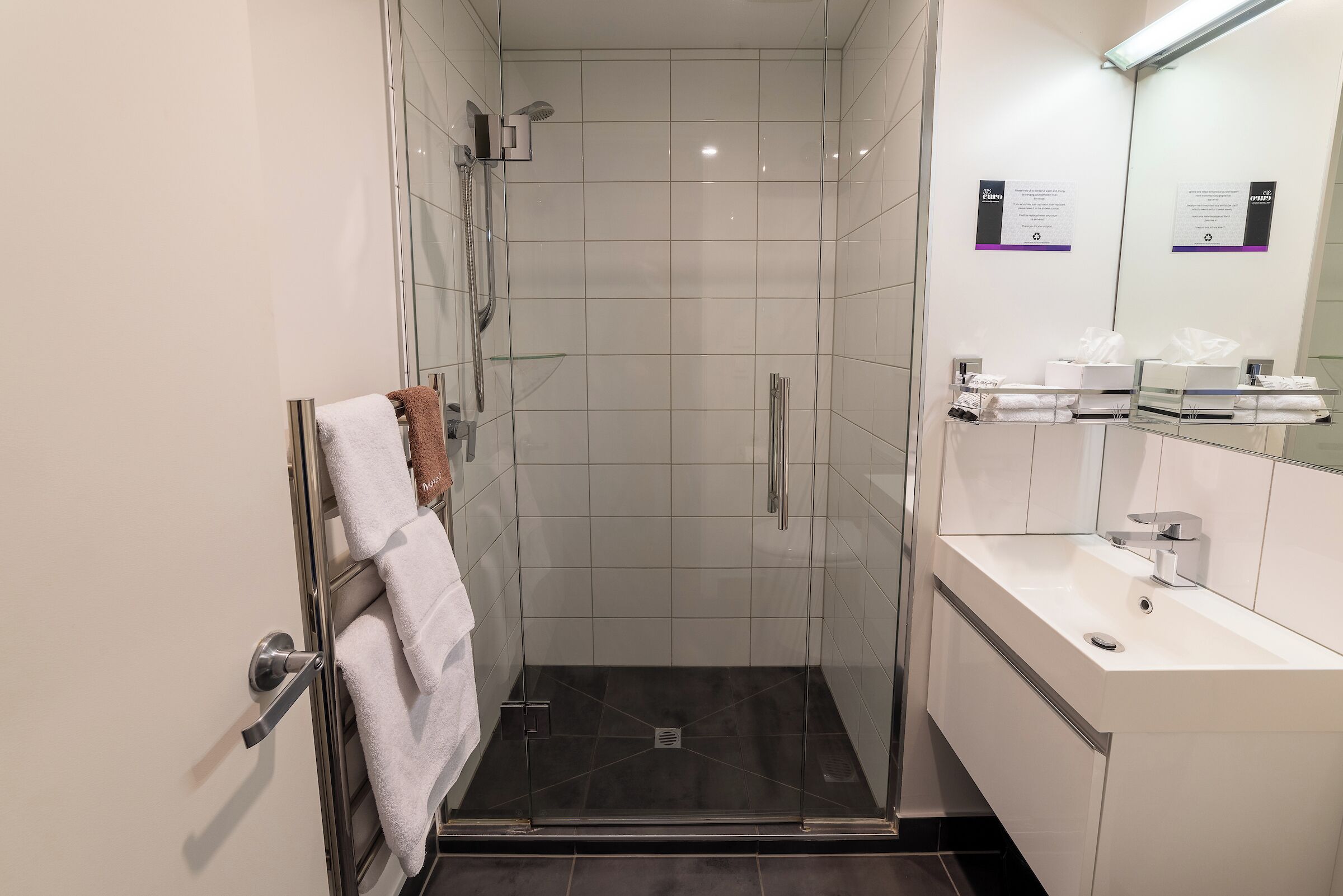 Un baño moderno con ducha de vidrio, lavamanos blanco y un espejo con un cartel.
