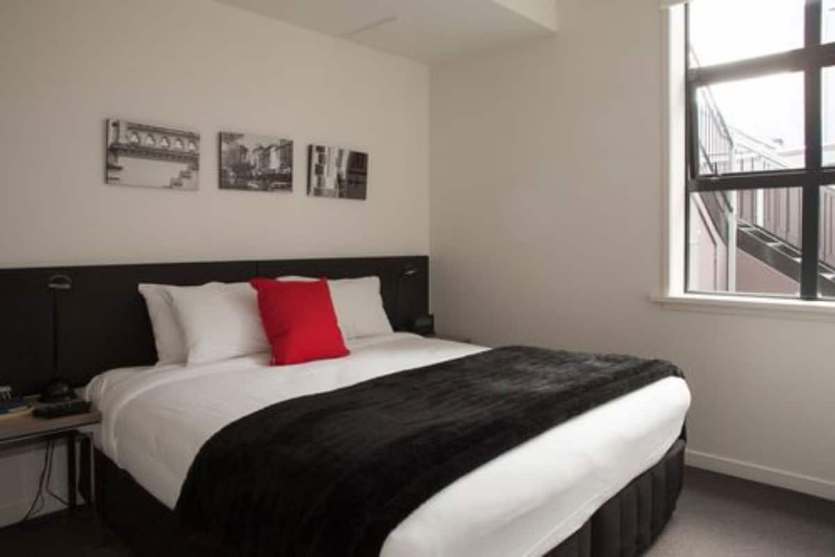 A hotel room with a large bed, a black headboard, a window with a view of a building, and three framed pictures on the wall.