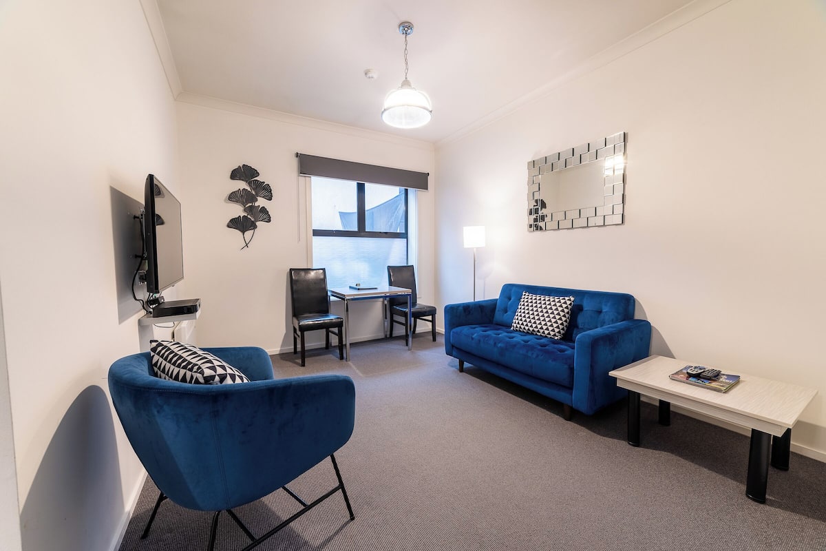 A modern living room with a blue sofa, a small table, a TV mounted on the wall, and a window with a view.