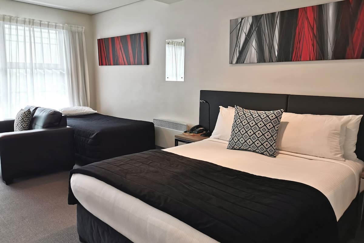 A hotel room with a large bed, two smaller beds, a desk, and a chair. There are two paintings on the wall and a window with curtains.