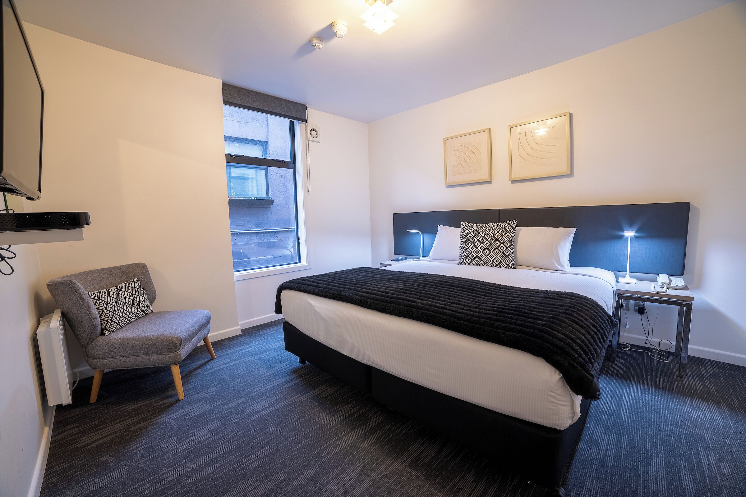 A modern hotel room with a large bed, a bedside table with a lamp, a chair, and a window with blinds.