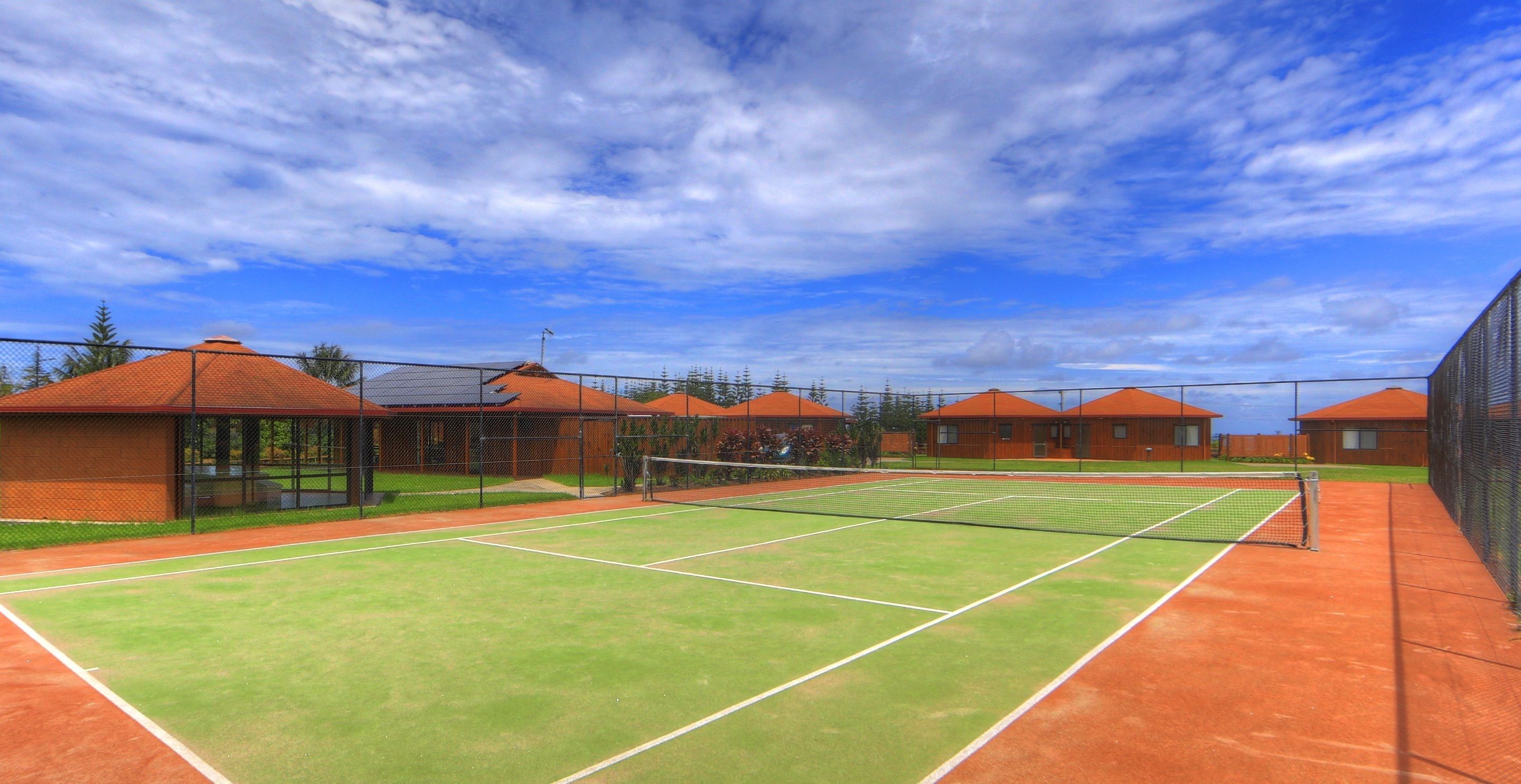 A tennis court with a net, surrounded by buildings with red roofs and a clear sky.
