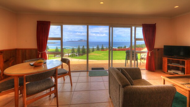 A room with a large sliding glass door leading to a balcony with a view of the ocean. Inside, there is a round table with chairs, a sofa, and a television on a wooden stand. - Islander Lodge Apartments (Norfolk Island)