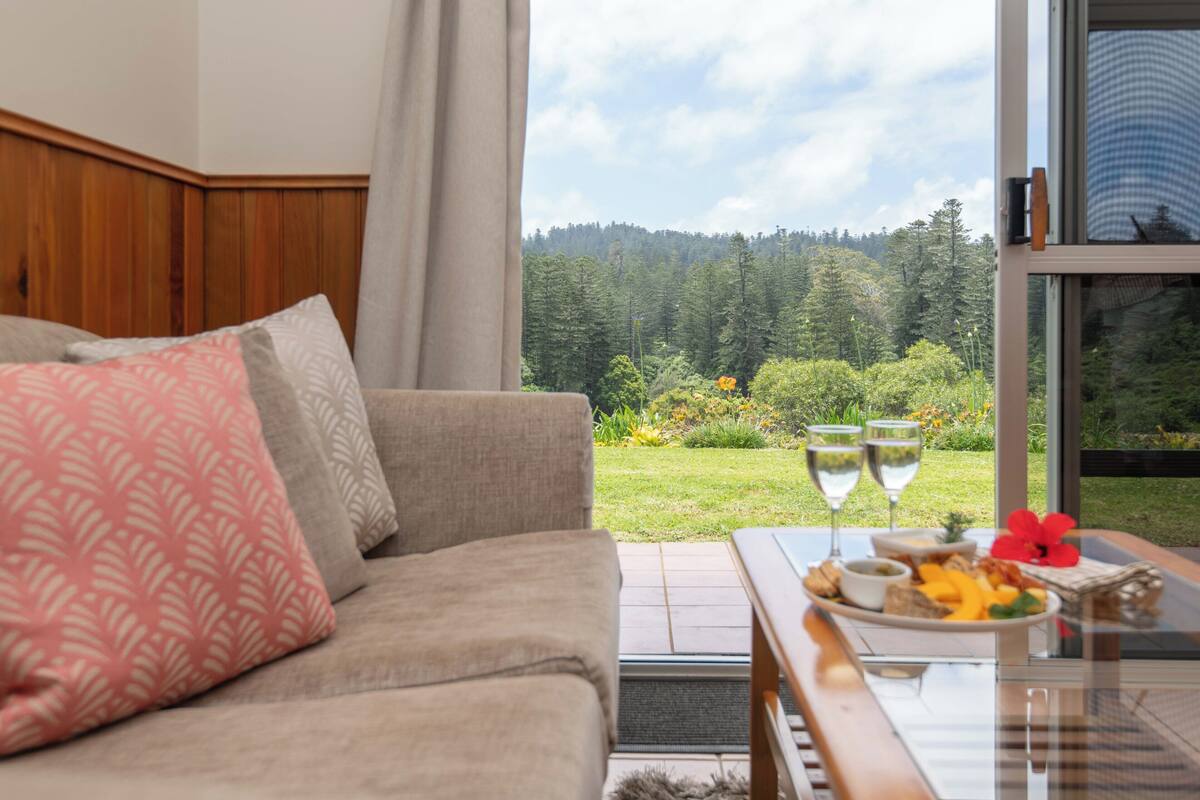 A living room with a sofa, a glass coffee table with a plate of food and two wine glasses, and a view of a forested area through a window.