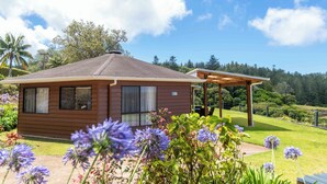 A small wooden house with a covered patio, surrounded by lush greenery and purple flowers. - Whispering Pines (Norfolk Island)