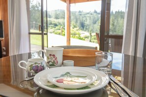 A table set with a plate, a cup, a glass, and a bowl, with a scenic view through a window.