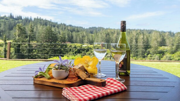 A wooden table with a bottle of wine, two glasses, a bowl of fruit, and a checkered napkin. - Whispering Pines (Norfolk Island)