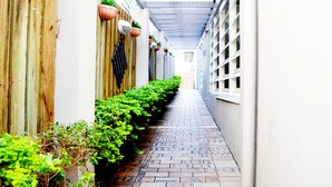 Uma estreita passagem de tijolos ladeada por cercas de madeira e plantas em vasos, que conduz a um edifício com fachada branca e uma galeria coberta.