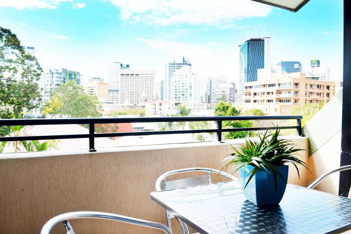 A balcony with a table and chairs, a potted plant, and a city skyline in the background.