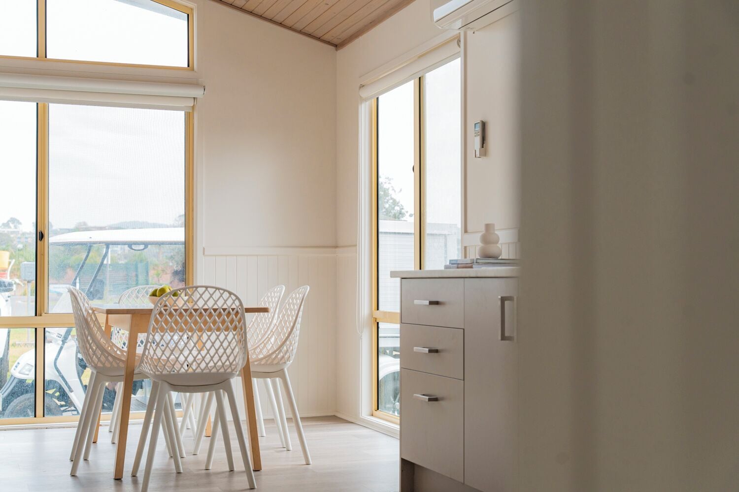 A modern kitchen with white cabinetry, a dining table with chairs, and large windows offering a view of the outdoors.