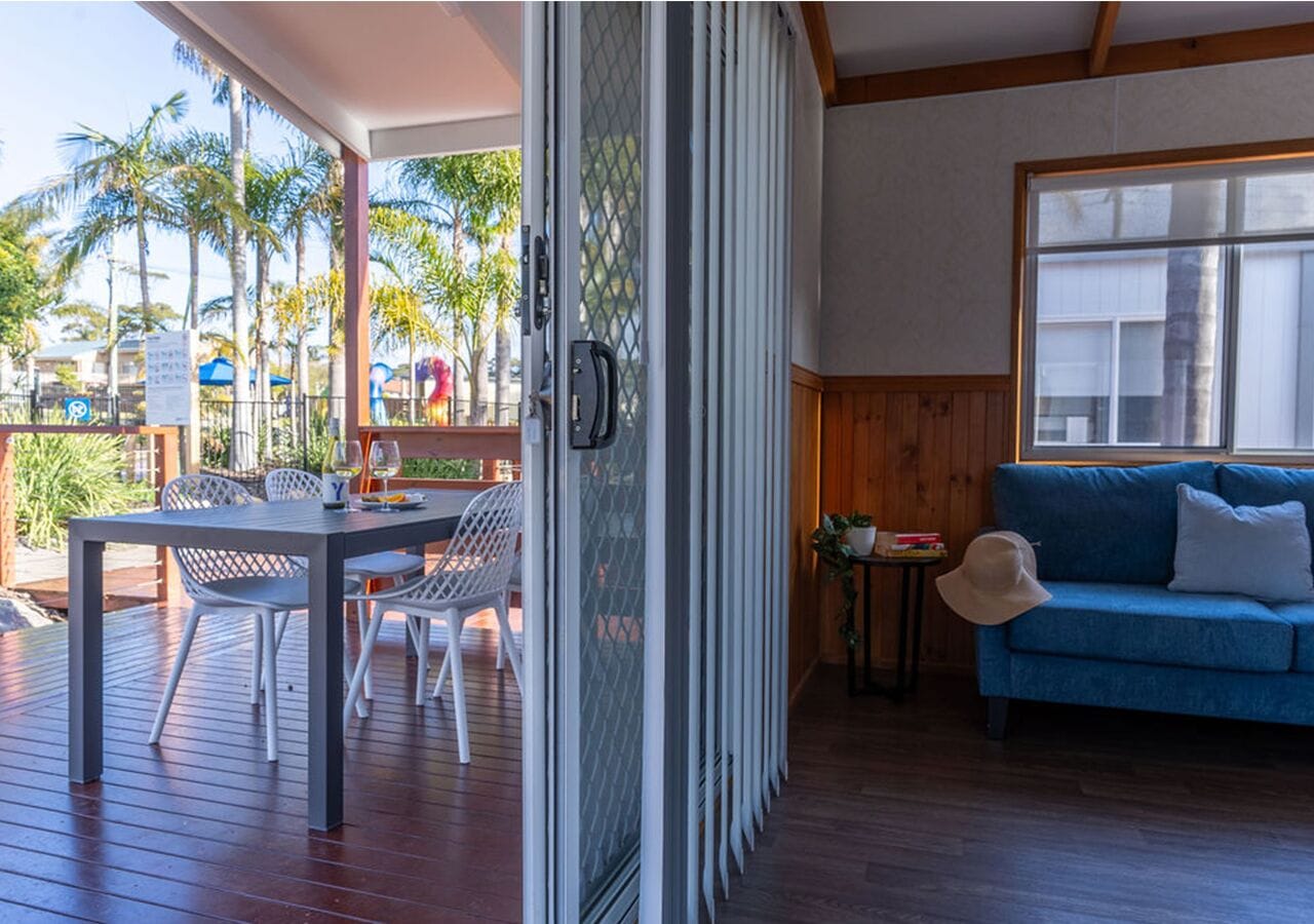 A room with a wooden deck, a dining table with chairs, a blue sofa, and a sliding glass door leading to an outdoor area with palm trees.
