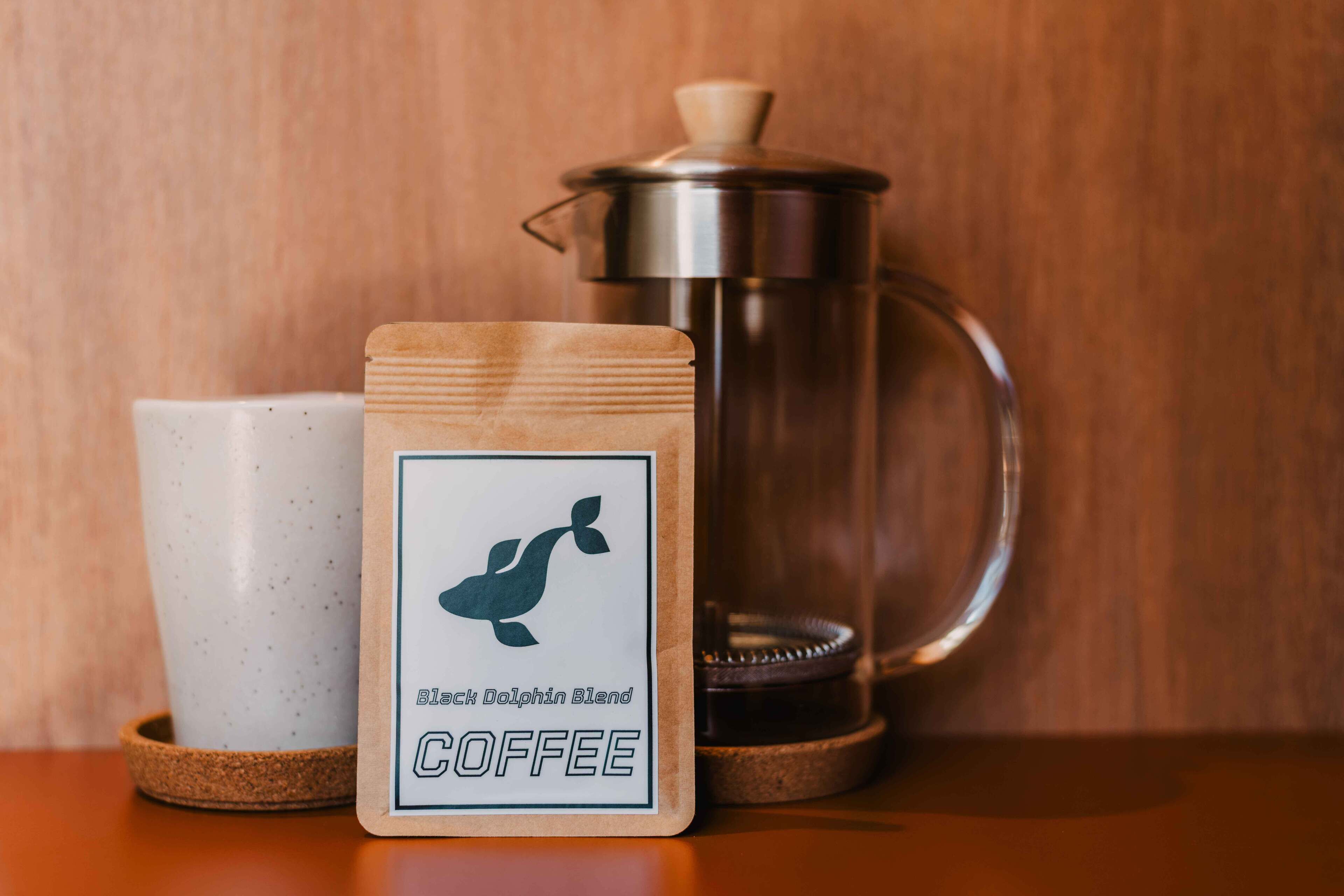 A coffee maker, a coffee pod labeled "Black Dolphin Blend Coffee," and a white mug on a wooden surface.