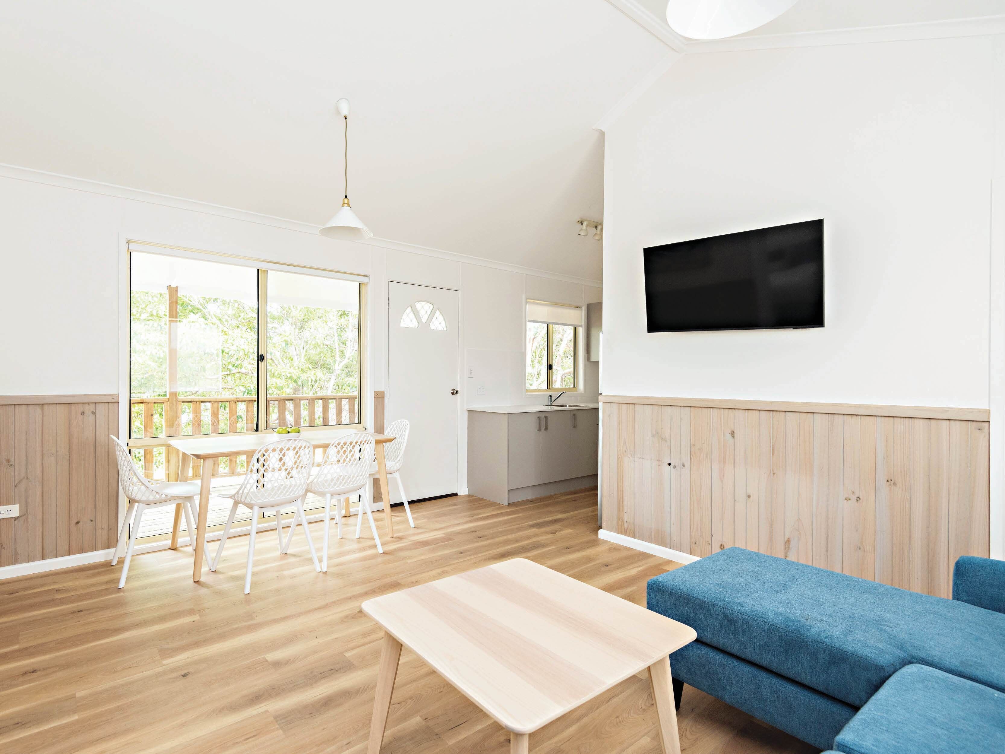 A spacious living area with a wooden floor, a dining table with chairs, a blue sofa, and a TV mounted on the wall.