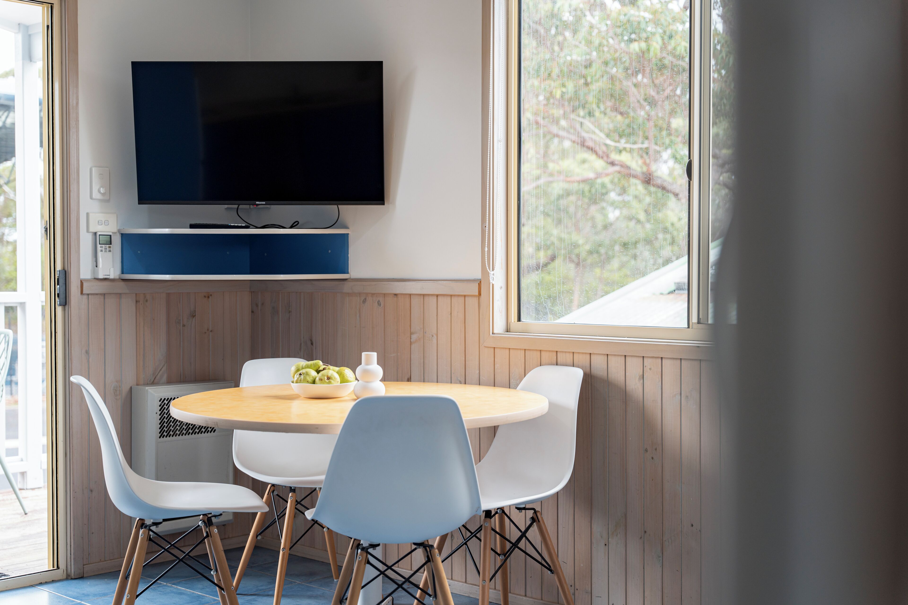 Un comedor con una mesa redonda, sillas blancas y azules, un televisor de pantalla plana en la pared y una ventana con vistas a árboles.
