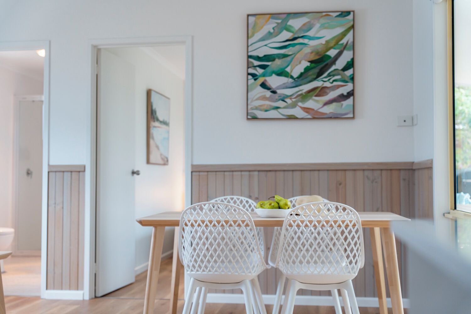 A dining area with a wooden table and white chairs, a painting on the wall, and a door leading to another room.