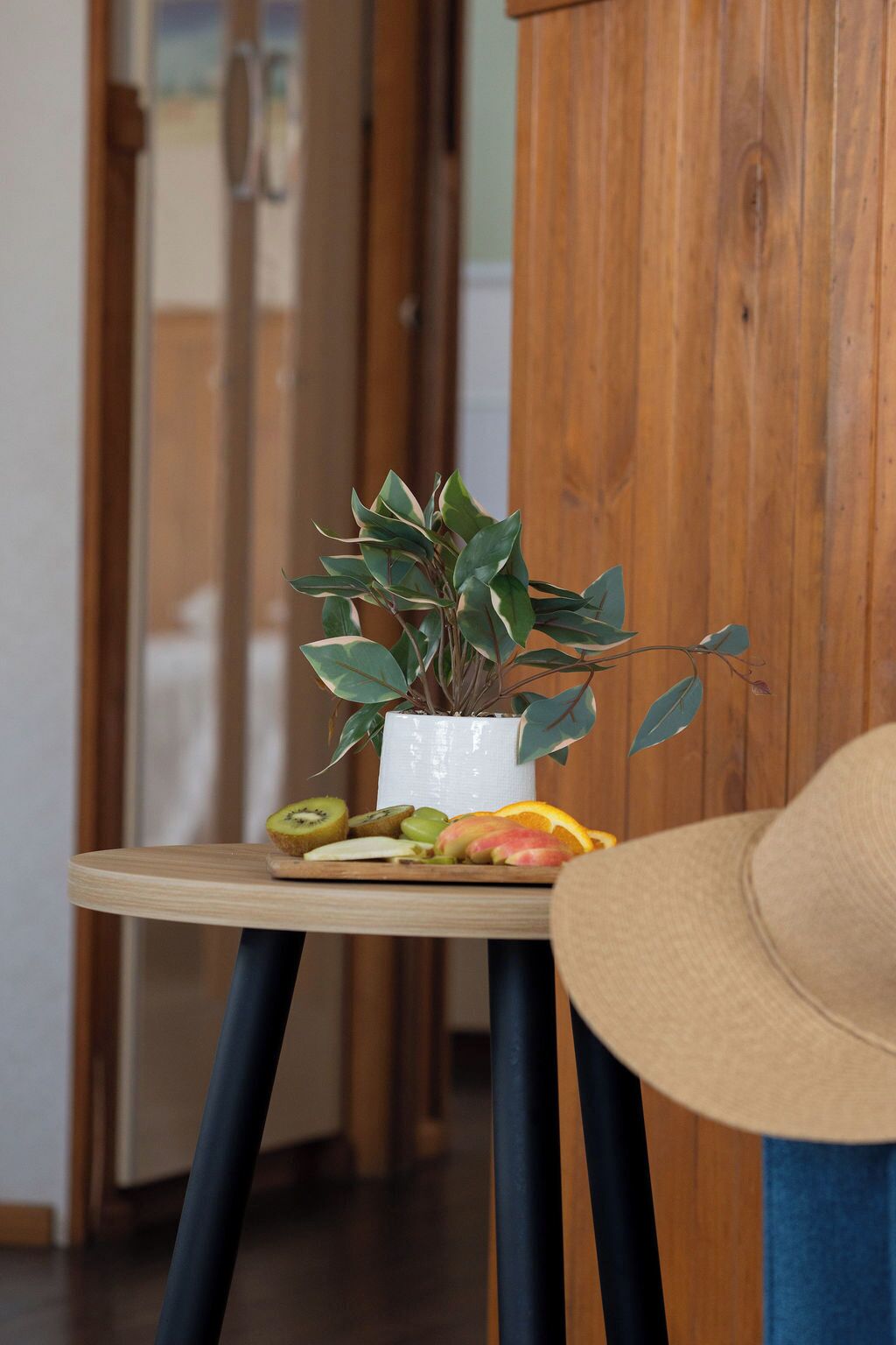 A wooden chair with a hat on it, a small table with a potted plant and fruit, and a wooden cabinet in the background.