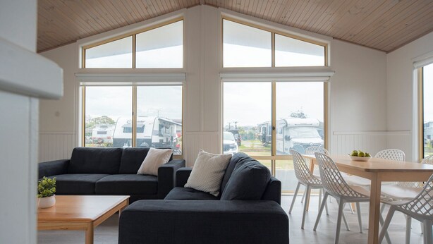 A modern living room with a dark grey sectional sofa, a wooden coffee table, and a dining area with white chairs and a wooden table. Large windows provide a view of the outside. - The Black Dolphin Holiday Park (Merimbula)