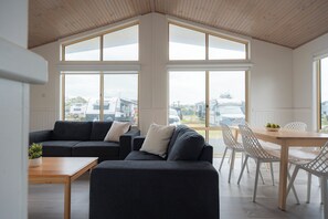 A modern living room with a dark grey sectional sofa, a wooden coffee table, and a dining area with white chairs and a wooden table. Large windows provide a view of the outside. - The Black Dolphin Holiday Park (Merimbula)
