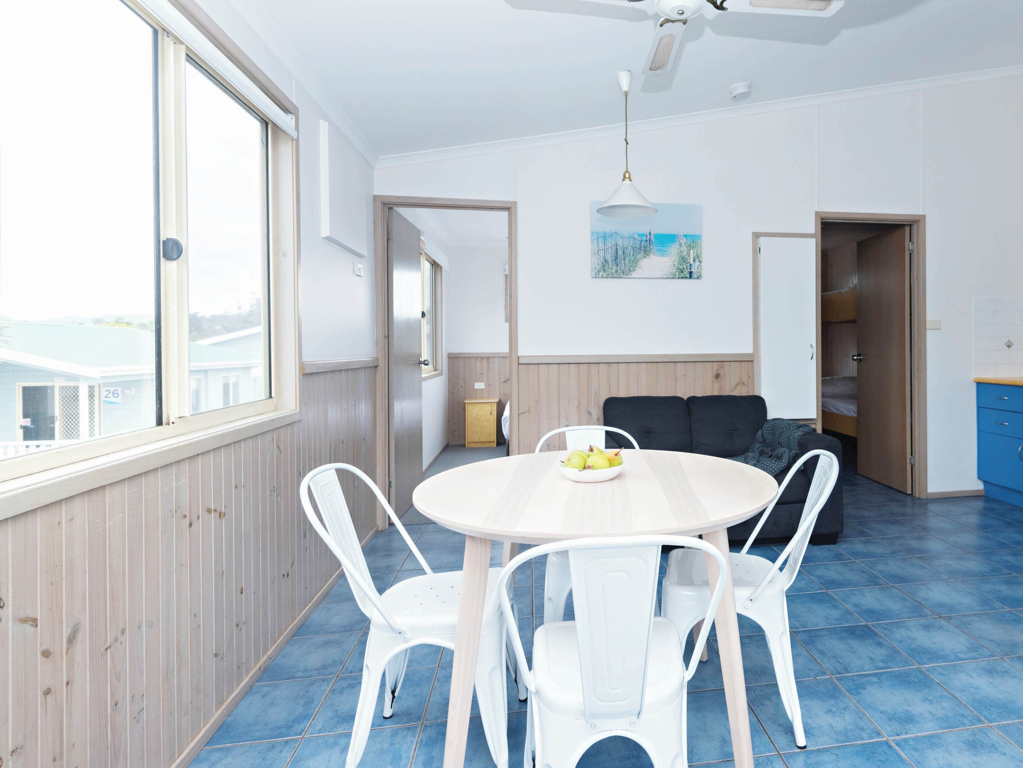 A room with a round table and white chairs, a sofa, a painting on the wall, and a view through a window.