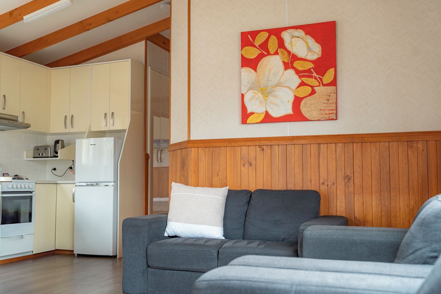 A living room with a grey sofa, a white pillow, wooden paneling on the wall, and a kitchen in the background.