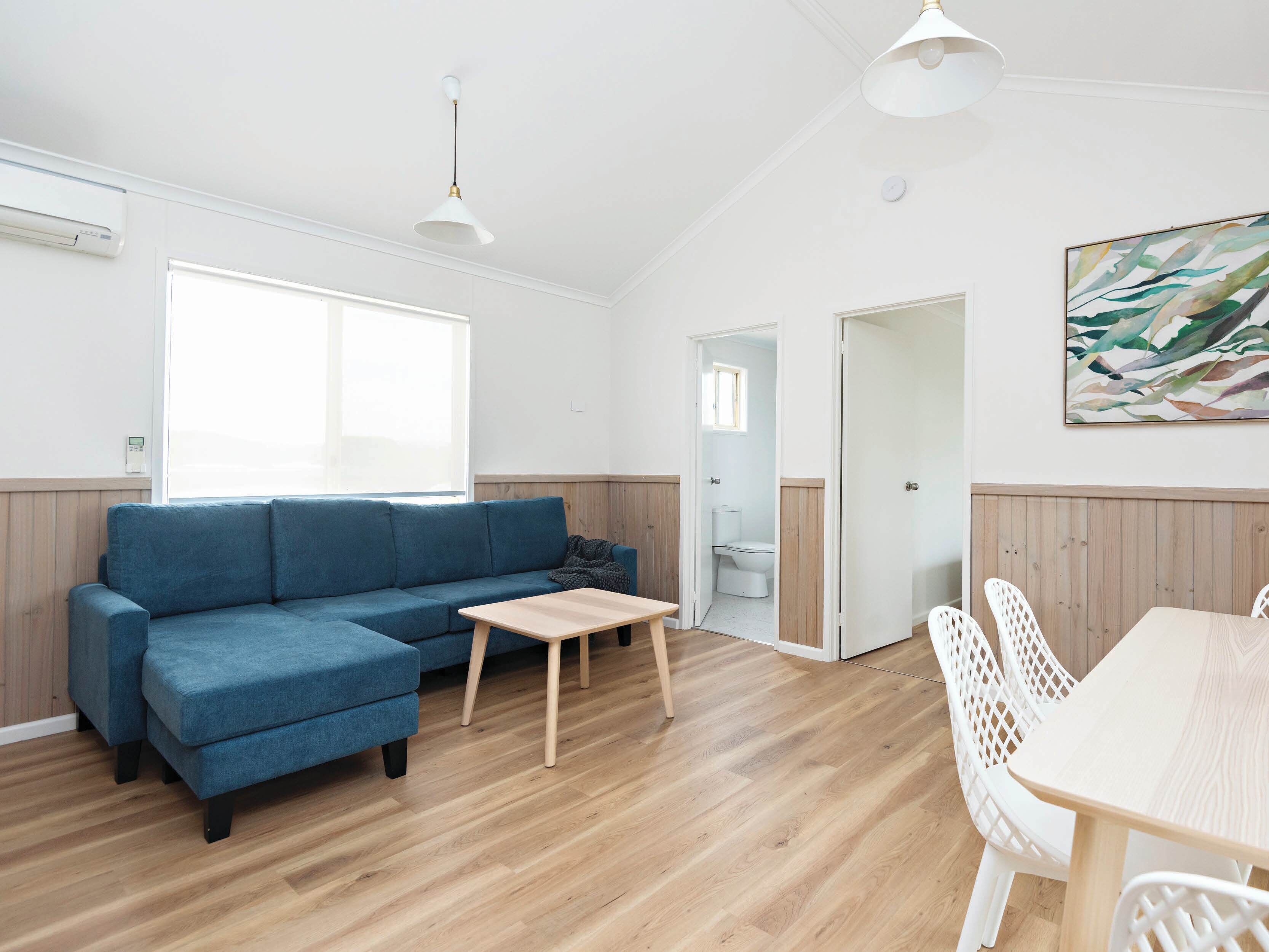 A modern living room with a blue sofa, a wooden coffee table, and a dining area with white chairs.